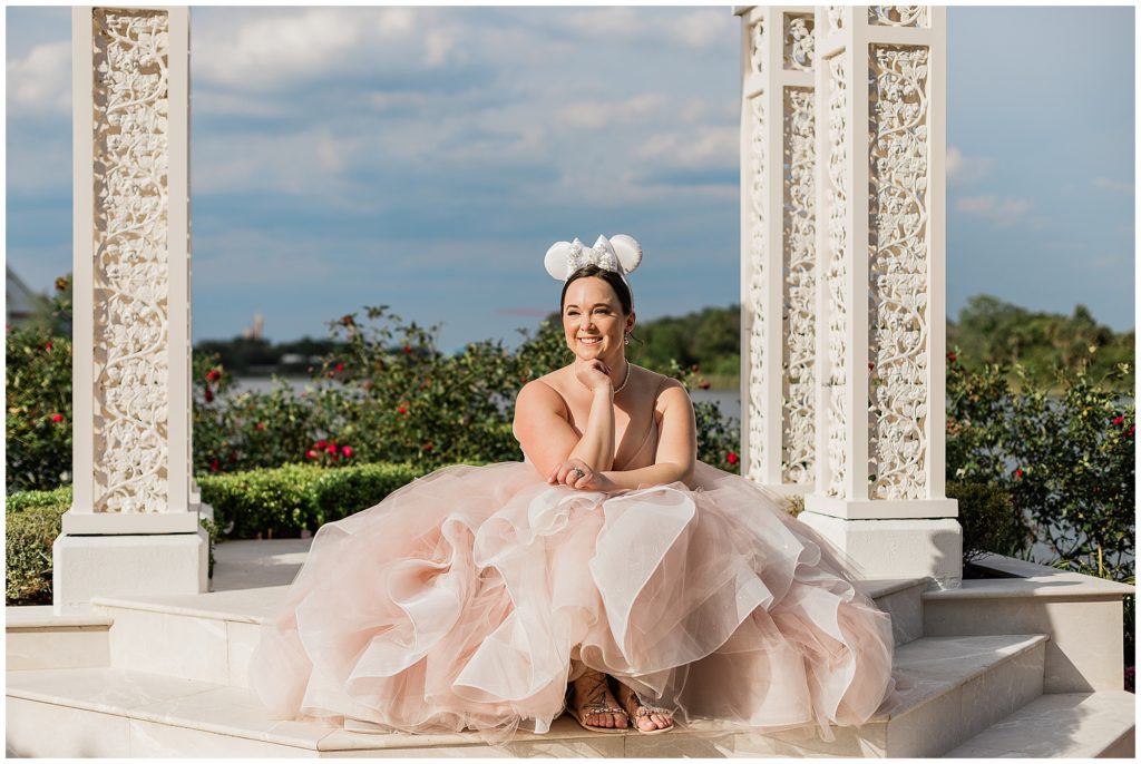 Bride sitting down with chin on hand smiling wearing pink gown and Minnie ears at Picture Point 